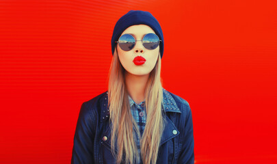 Wall Mural - Portrait of stylish beautiful blonde woman blowing her red lips sending sweet air kiss wearing round sunglasses, black rock style, jacket and hat