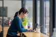 © amnaj - Young Asian Woman Working on Laptop in a Cozy Cafe, Freelancer at a Coffee Shop