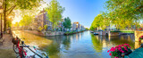 Fototapeta  - Soul of Amsterdam. Early morning in Amsterdam. Ancient houses, bridges, traditional bicycles, canals, boats,  and the sun shines through the trees. Panoramic view with all the sights of Amsterdam.