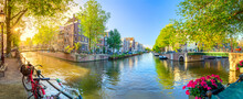 Soul Of Amsterdam. Early Morning In Amsterdam. Ancient Houses, Bridges, Traditional Bicycles, Canals, Boats,  And The Sun Shines Through The Trees. Panoramic View With All The Sights Of Amsterdam.
