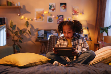 Full length portrait of teenage black girl with pigtails using digital tablet while sitting on bed at home in cozy warm light, copy space