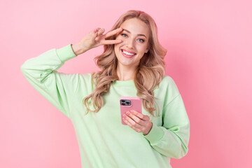 Poster - Portrait of young girlfriend showing v sign over her face blogging influencer wear green trendy pullover isolated on pink color background