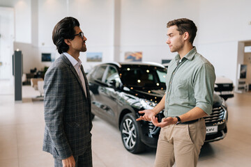 Wall Mural - Side view of handsome male client talking to car dealer wearing business suit in dealership discussing automobiles looking at luxurious new model. Concept of choosing and buying new auto at showroom.