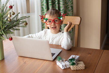 Little boy in a white sweater and funny glasses sits at a laptop and writes a letter to Santa Claus