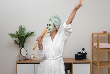 Happy woman with clay mask on face singing in hairbrush mic and dancing at home lady in bathrobe with towel on head having fun on pampering day skincare treatment