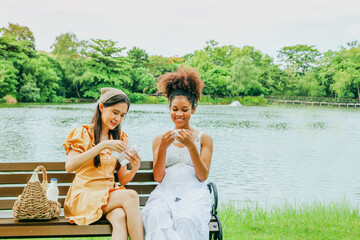 Wall Mural - Cute asian woman carries fruit basket and takes her healthy pregnant african american female friend picnic beautiful garden shady atmosphere sitting wooden bench lovingly giving gift of baby socks.