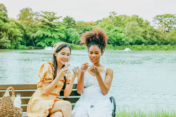 Wall Mural - Cute asian woman carries fruit basket and takes her healthy pregnant african american female friend picnic beautiful garden shady atmosphere sitting wooden bench lovingly giving gift of baby socks.