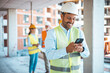 © Dragana Gordic - Shot of a young businessman using his smartphone while on a construction site. Portrait of young engineer in vest with white helmet standing on construction site, smiling and holding smartphone