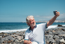 Happy White-haired Senior Man Enjoying Sea Vacation Sitting At The Beach In A Sunny Day Taking A Selfie With Mobile Phone