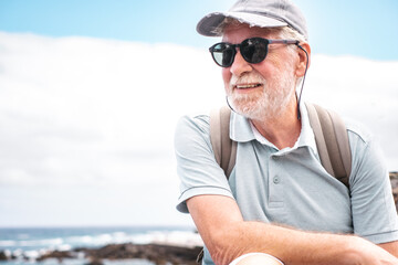 Portrait of smiling bearded senior man with cap and sunglasses sitting at sea carrying backpack enjoying summer vacation