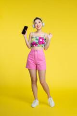 Wall Mural - Asian pretty woman wearing headphones and holding on her cell phone over yellow background.