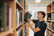 © Maskot - Side view of male student removing book from bookshelf in library at university