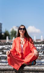 Wall Mural - Summer portrait of a girl with long blond hair. A girl in orange clothes and sunglasses sits in a lotus position near the city fountain.