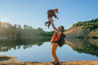 © Iona - Mid aged caucasian man throwing up in air his son against lake at sunset time enjoying summer sunset outdoors, tourism, family leisure. Father  and son with backpacks at nature. Travel, togetherness.