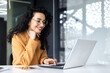 © Liubomir - Happy and smiling hispanic businesswoman typing on laptop, office worker with curly hair and glasses happy with achievement results, at work inside office building. .
