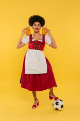 Wall Mural - cheerful african american waitress in dirndl standing with beer mugs near soccer ball on yellow