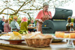 © Lomb - Woman at the BBQ with Friends in the Countryside - An elderly lady, the focal point, grills amidst a relaxed gathering, with soft-focus dishes in the foreground and cheerful friends in the background.