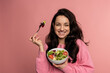 © Nataliya - Waist-up portrait of a pleased lady eating a portion of an appetizing vegetable salad with the fork from the bowl during the studio photo shoot..