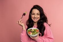 Waist-up Portrait Of A Pleased Lady Eating A Portion Of An Appetizing Vegetable Salad With The Fork From The Bowl During The Studio Photo Shoot..
