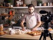 © The Little Hut - Generative AI : Baking process. Young cooking man speaker talking on digital camera recording vlog.