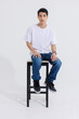 © Bangkok Click Studio - Portrait isolated cutout studio shot Asian handsome cool male fashion model in casual fashionable t shirt and blue jeans sitting on chair holding hand in pocket look at camera on white background