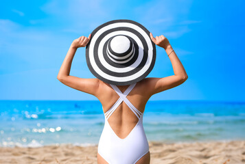 Wall Mural - A woman wearing a white swimsuit and wearing a black and white Hawaiian hat. She stands holding a Hawaiian hat. and looking at the beach and the sea She travels on vacation.