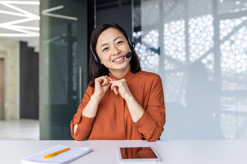 Webcam view, online video call remote meeting, businesswoman with headset phone smiling and looking at camera, asian woman talking with colleagues using laptop while sitting inside office.