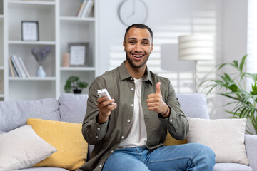 Portrait of a young African American man sitting on the sofa at home and holding the air conditioner remote control, smiling at the camera and showing the super gesture with his finger