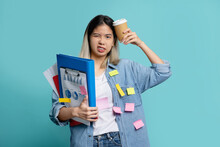 Bored Asian Female Office Worker With A Lot Of Work With Holding Document File  With Cup Of Coffee And Note On Face Standing On Blue Background In Studio.