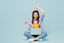 Full Body Young Happy IT Woman Wear Purple Shirt Yellow T-shirt Casual Clothes Sit Hold Use Work On Laptop Pc Computer Point Index Finger On Isolated On Plain Pastel Blue Background Studio Portrait.