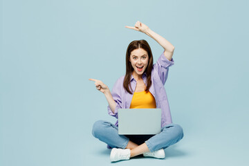 Full body young happy IT woman wear purple shirt yellow t-shirt casual clothes sit hold use work on laptop pc computer point index finger on isolated on plain pastel blue background studio portrait.