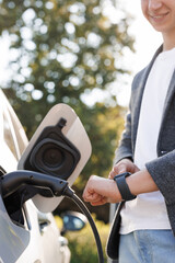 Unrecognizable man plugging in charging cable to to electric vehicle. Male hand inserts power connector into EV car and charges batteries uses smartwatch for activates start charging