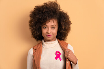 Wall Mural - confident black young woman showing pink ribbon in beige background. breast cancer, awareness concept.