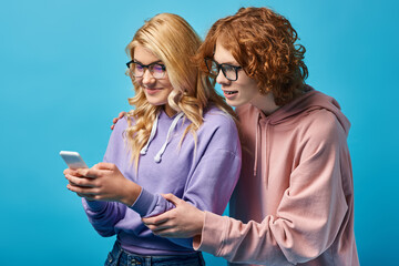 Wall Mural - joyful blonde teen girl in eyeglasses and trendy hoodie using smartphone near redhead friend on blue