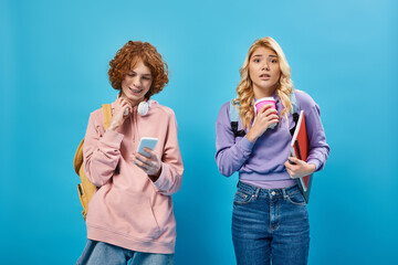 Wall Mural - nervous teen girl with paper cup looking at camera near positive friend using smartphone on blue