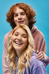 Wall Mural - portrait of carefree teenage friends, redhead guy and blonde girlfriend looking at camera on blue