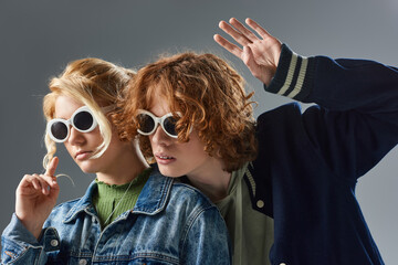 Wall Mural - redhead teenage guy waving hand near blonde girl in casual clothes and trendy sunglasses on grey