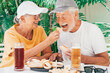 Handsome cheerful senior couple in hats sitting at pub restaurant table eating sandwiches on a sunny summer day while enjoying glasses of cold beer