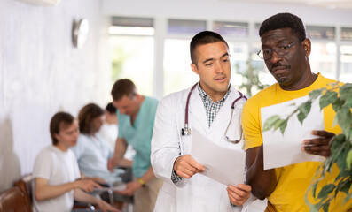 Wall Mural - Young male doctor discussing medical document with concerned middle-aged patient standing in waiting room