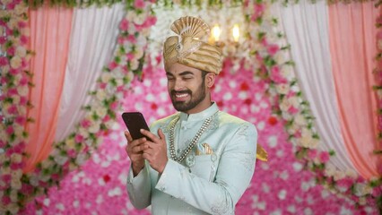 Wall Mural - Happy smiling young man as Groom or wedding ethnic wear using mobil phone on decorated background - concept of social media sharing, technology and conversation