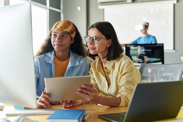 Young teacher using digital tablet and working with student at lesson while they sitting at table with computer