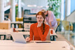 © .shock - In a modern startup office, a professional businesswoman with orange hair sitting at her laptop, epitomizing innovation and productivity in her contemporary workspace.