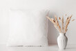 © Candle photo - Square white pillow stands near wall with dried flowers in vase. Mockup pillow close up in neutral color, minimalist interior.