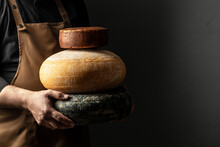 Craftsmanship Hands Have A Typical Italian Cheese. French Tomme Cheese In The Hands Of A Cheesemaker On Dark Background. Long Banner Format