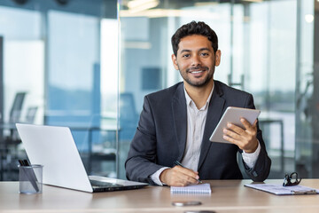 Portrait of young businessman with tablet computer inside office, hispanic man smiling and looking at camera, working with laptop, using app on tablet.