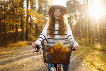 Happy Active Woman In Stylish Clothes Rides A Bicycle In An Autumn Park At Sunset. Outdoor Portrait. Beautiful Woman Enjoying Nature. Active Lifestyle.
