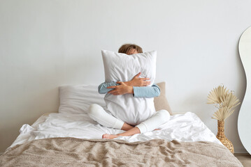 Time to sleep, sleep deficiency, good morning concept. Teenager boy sitting on bed, embracing white pillow, hiding face behind. Minimalist scandinavian neutral bedroom interior, lifestyle