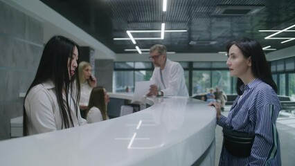 Wall Mural - Asian female administrator talks with patient at reception desk in clinic, gives terminal. Woman pays with contactless payment for appointment using watch. Medical stuff work in modern medical center.