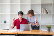 © PHAISITSAWAN - Cheerful business people using a laptop in an office. Happy young entrepreneurs smiling while working together in a modern workspace. Two young business people are sitting together at a table.
