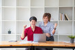 © PHAISITSAWAN - Cheerful business people using a laptop in an office. Happy young entrepreneurs smiling while working together in a modern workspace. Two young business people are sitting together at a table.